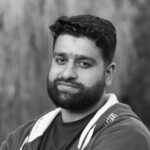 A serious man with a beard in a hoodie poses outdoors. Black and white portrait.