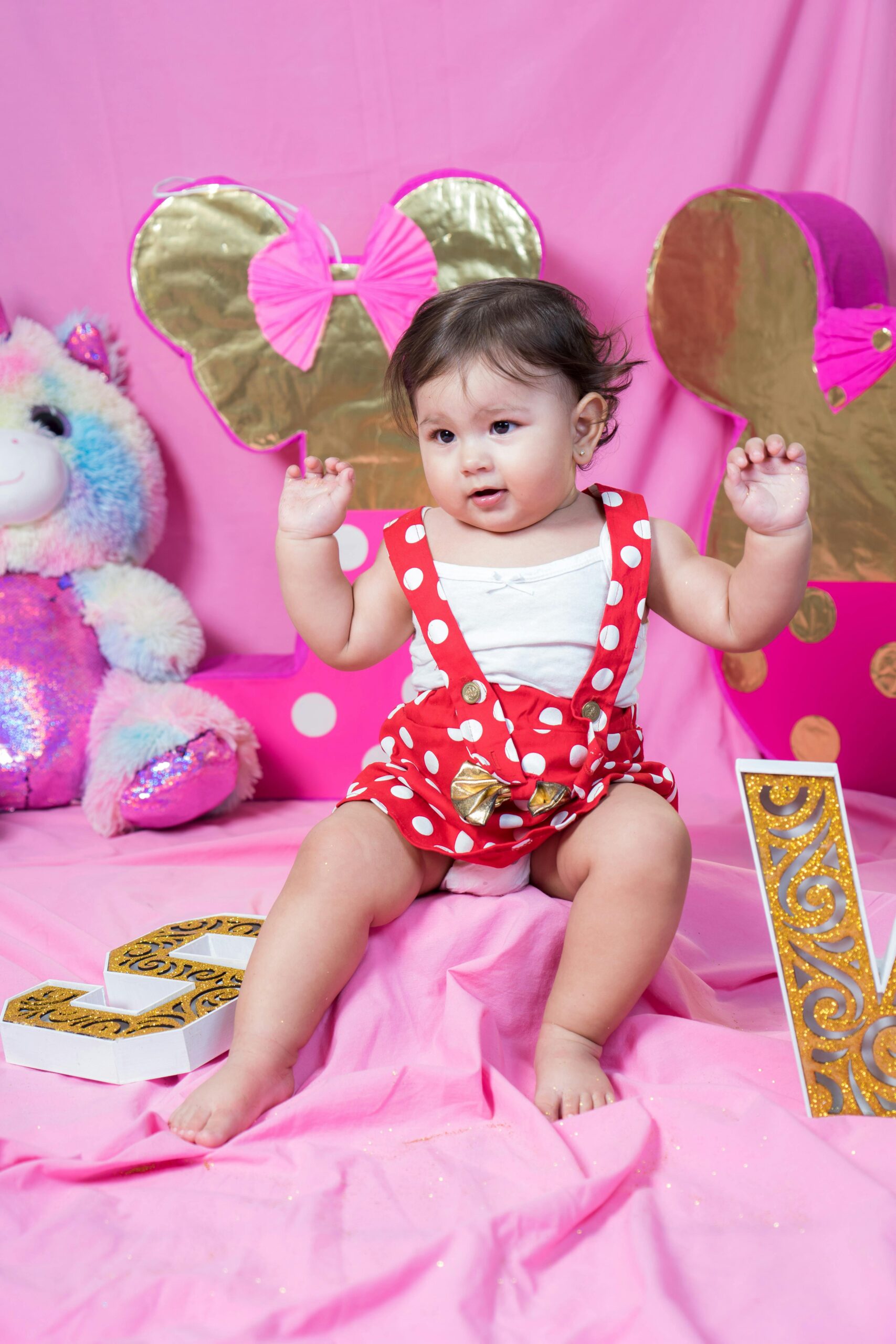 Cute baby girl in red polka dot outfit with pink birthday decorations and plush unicorn.
