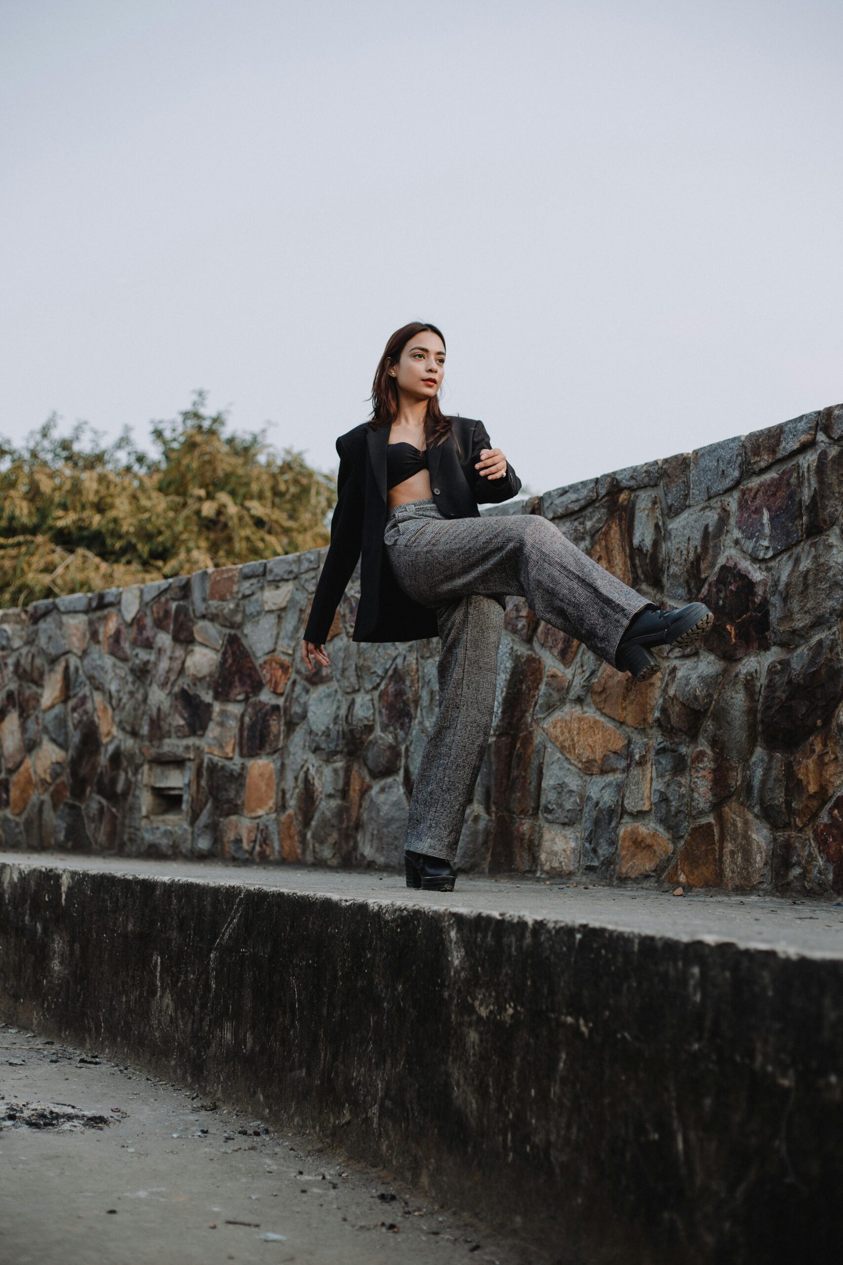 Confident woman in a blazer posing against a stone wall, exuding elegance and style in a natural setting.
